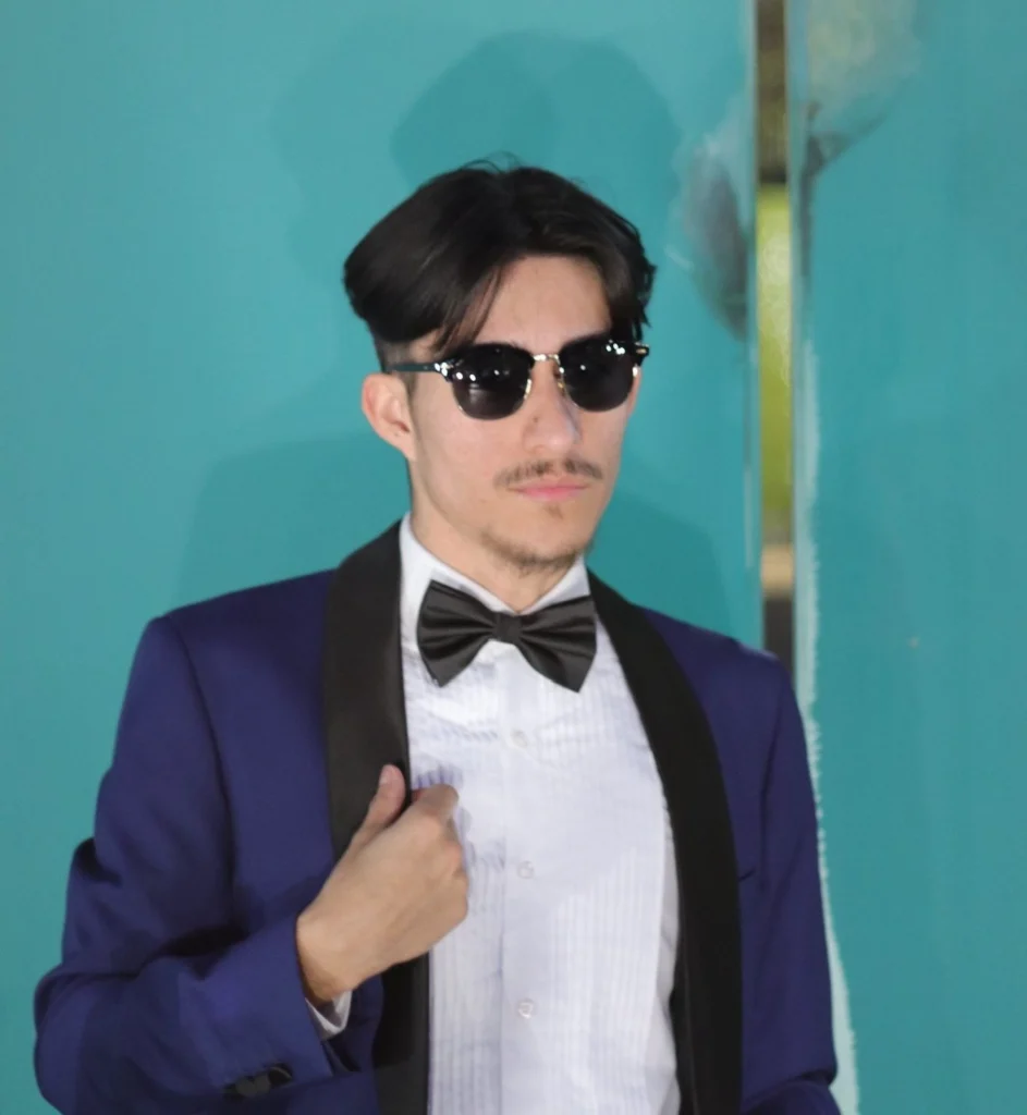 Man Wearing A Navy Blue Tuxedo With Black Bow Tie And Pleated Shirt Against A Teal Wall In Phoenix.