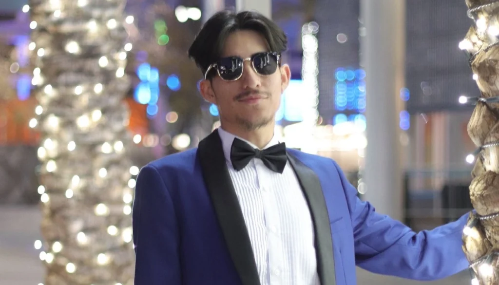 Man Wearing A Navy Blue Tuxedo With Black Bow Tie And Satin Lapels Standing Next To Palm Trees With String Lights At An Evening Event In Phoenix.