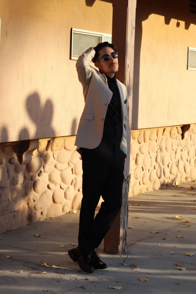 Full Body Shot Of A Man Wearing A Beige Suit Jacket, Black Shirt, Black Trousers, And Black Dress Shoes Leaning Against A Rustic Post In Phoenix.